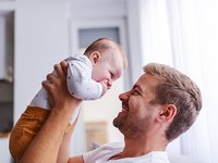 Junger Vater hebt Baby hoch, beide lächeln sich an