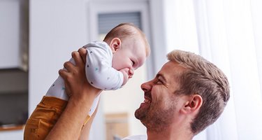 Junger Vater hebt Baby hoch, beide lächeln sich an